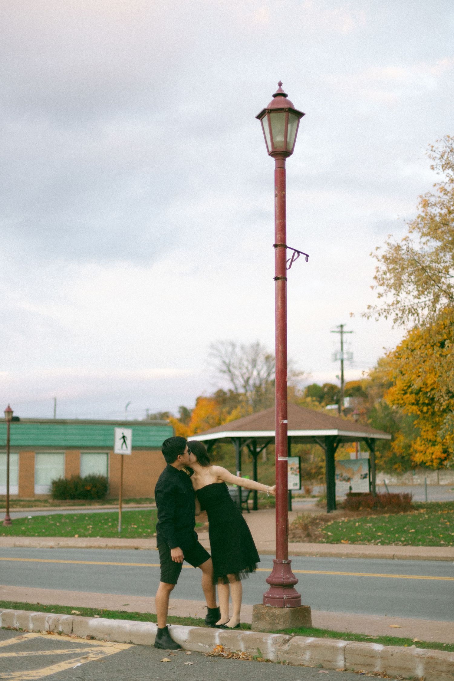 Two people in dark outfits stand together near a vintage red lamp post on a street with fall foliage in the background.