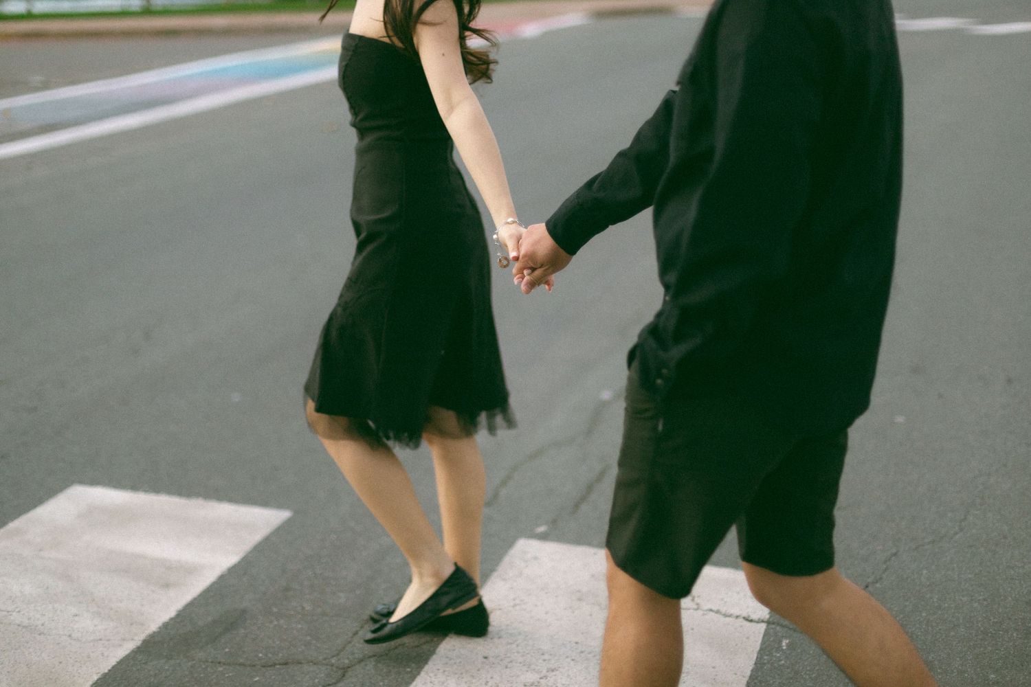 A couple holding hands while walking across a pedestrian crossing, with the woman wearing a black dress and flats.