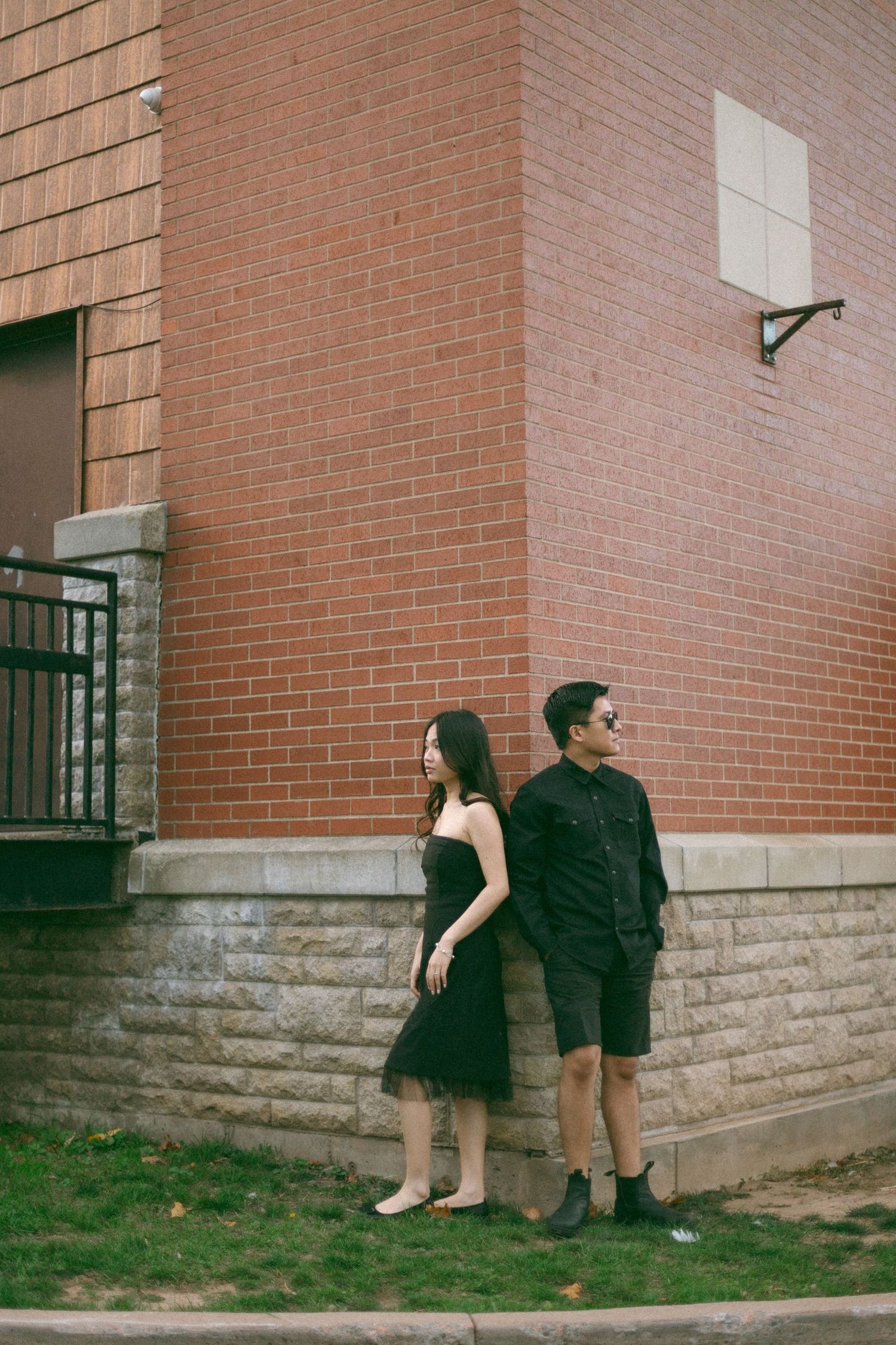A couple stands against a brick building corner, with her in a black dress and him in dark clothing.