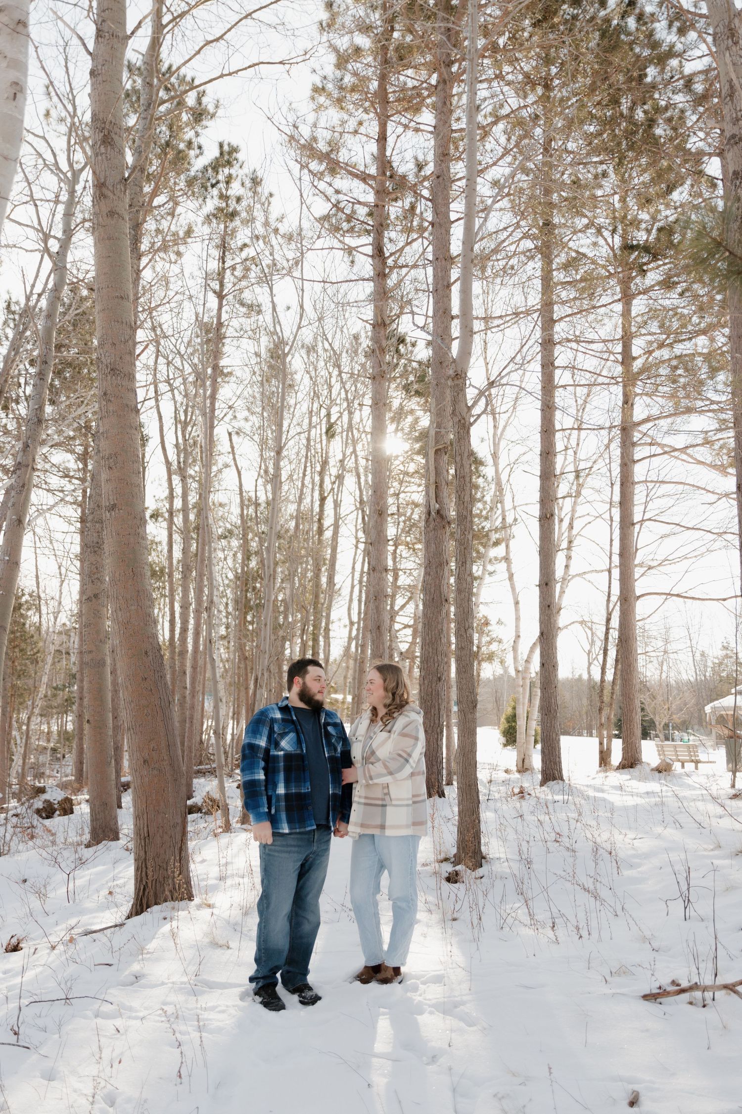 Two people walking together on a snow-covered trail through tall pine trees in winter.