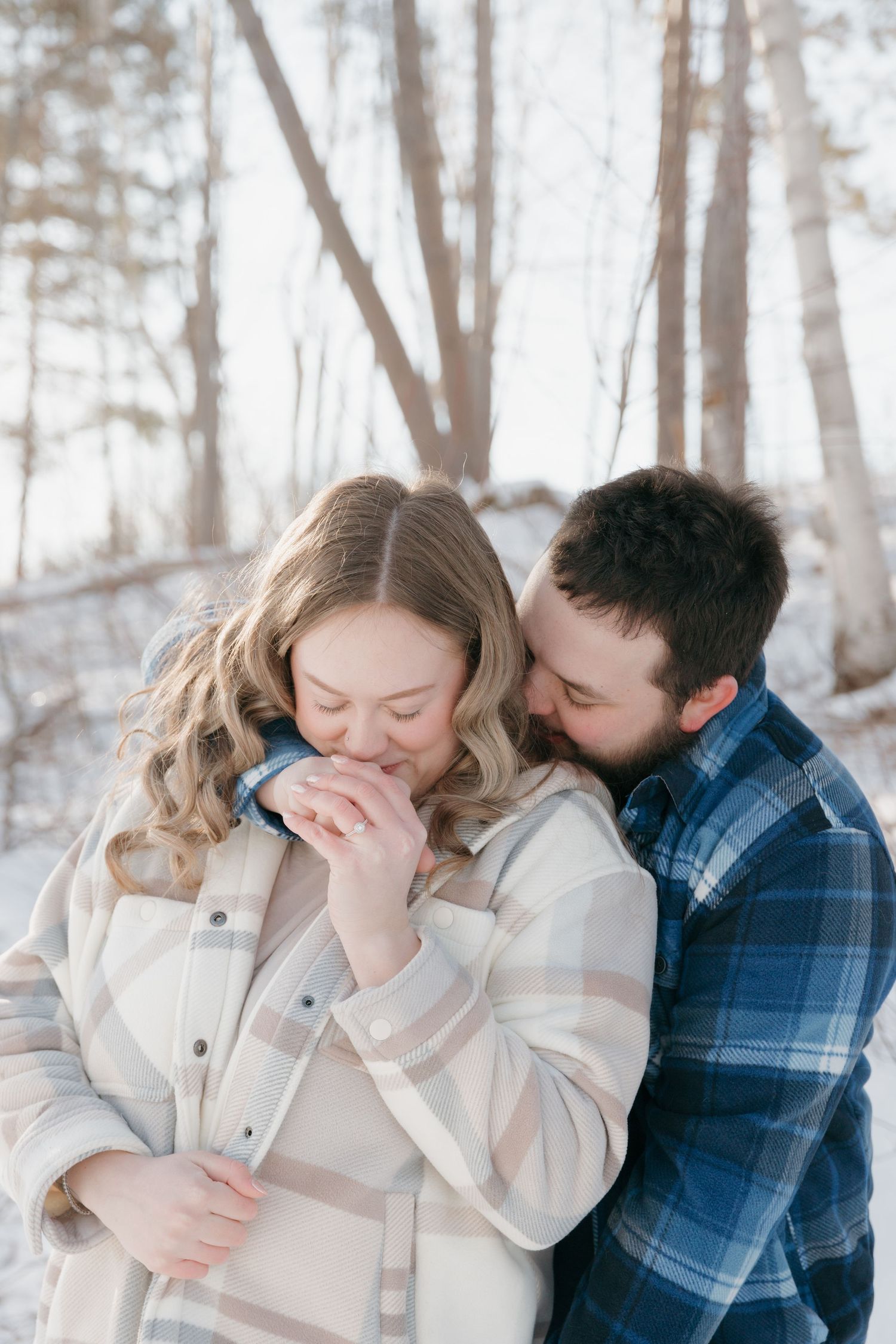 A couple shares an intimate winter moment in a snowy forest setting, dressed in a cream coat and plaid flannel.