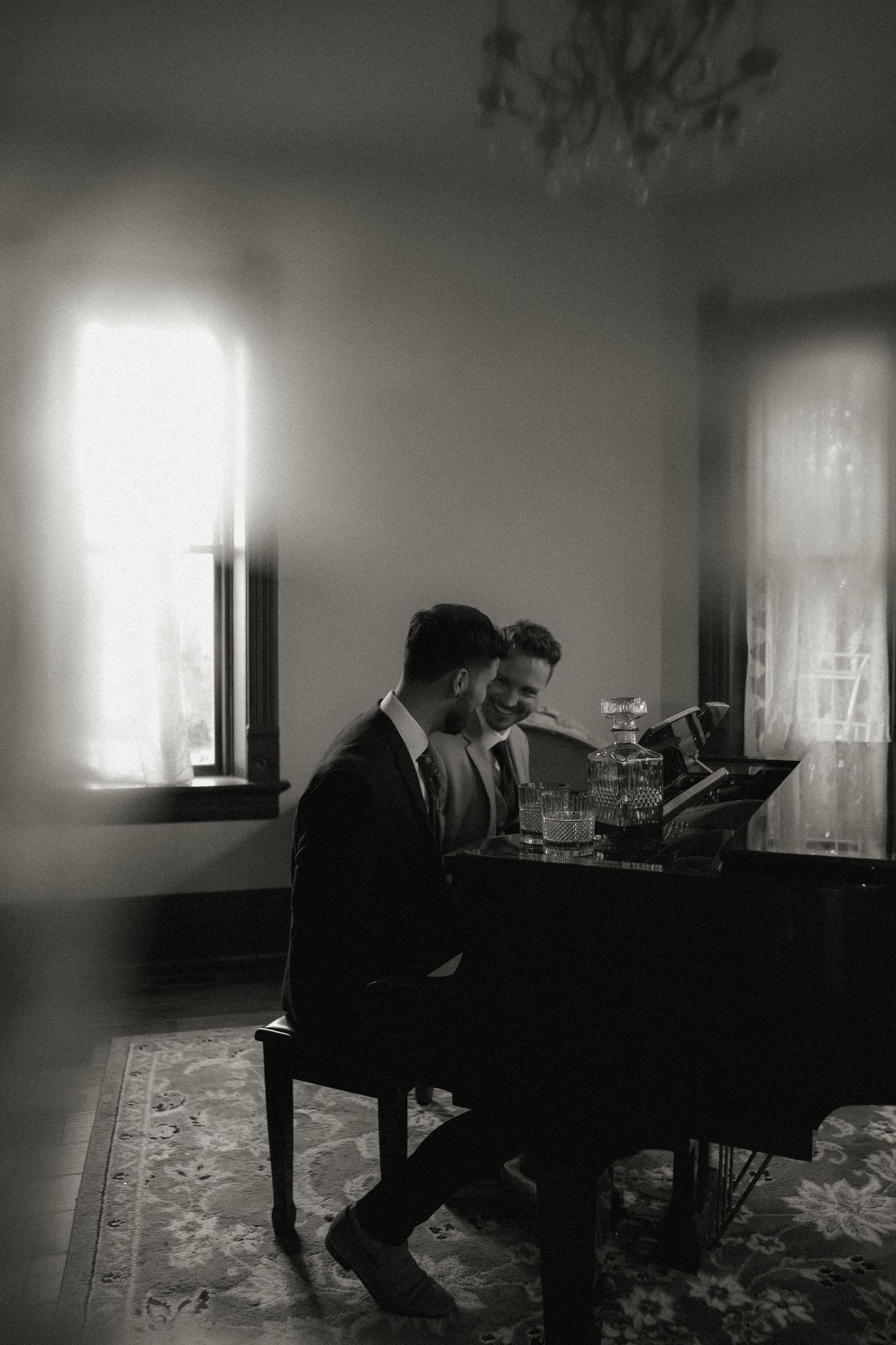 Two people seated at grand piano in dimly lit room with chandelier.