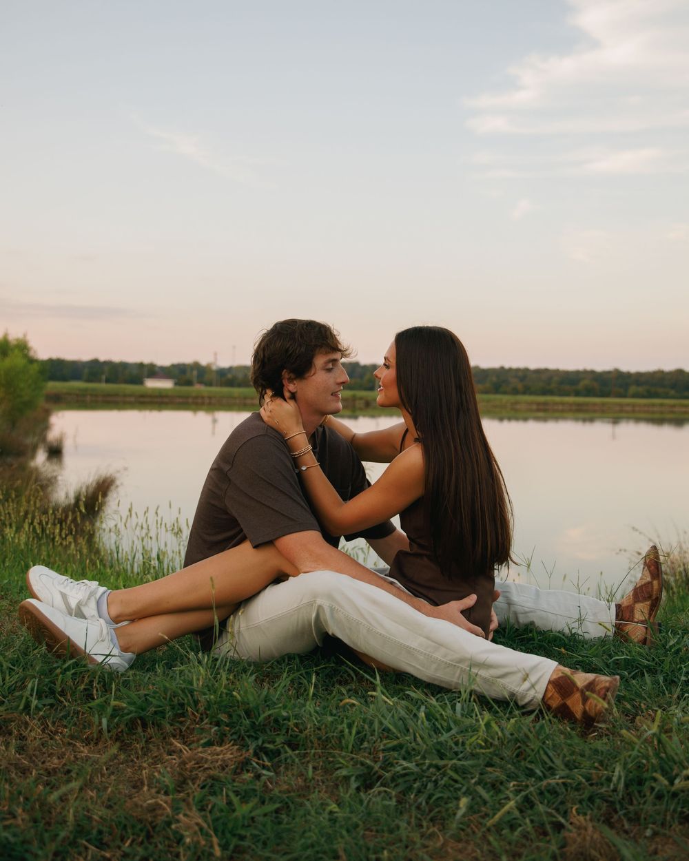 Romantic couple sitting on grass by a serene lake at sunset, embracing and gazing at each other.