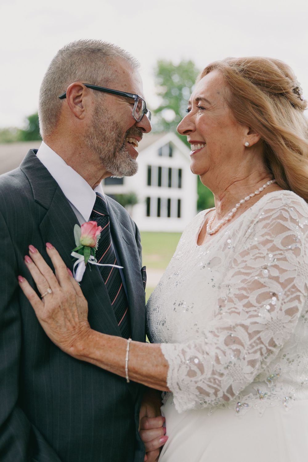 Older couple in wedding attire smiling at each other outdoors, groom in suit with boutonniere, bride in lace dress with pearls.