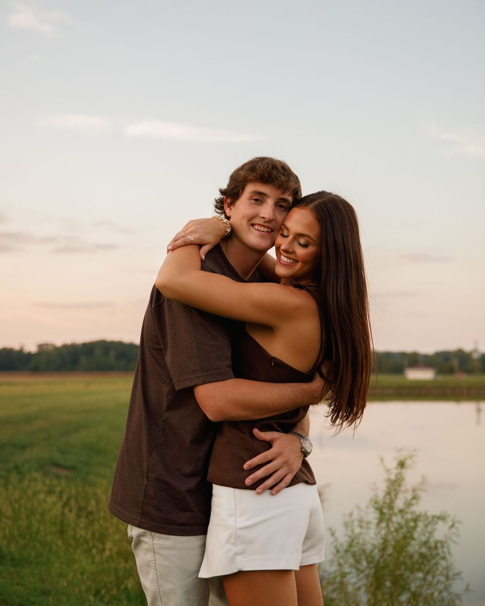 A smiling couple embraces outdoors near a lake at sunset, with green fields and trees in the background.
