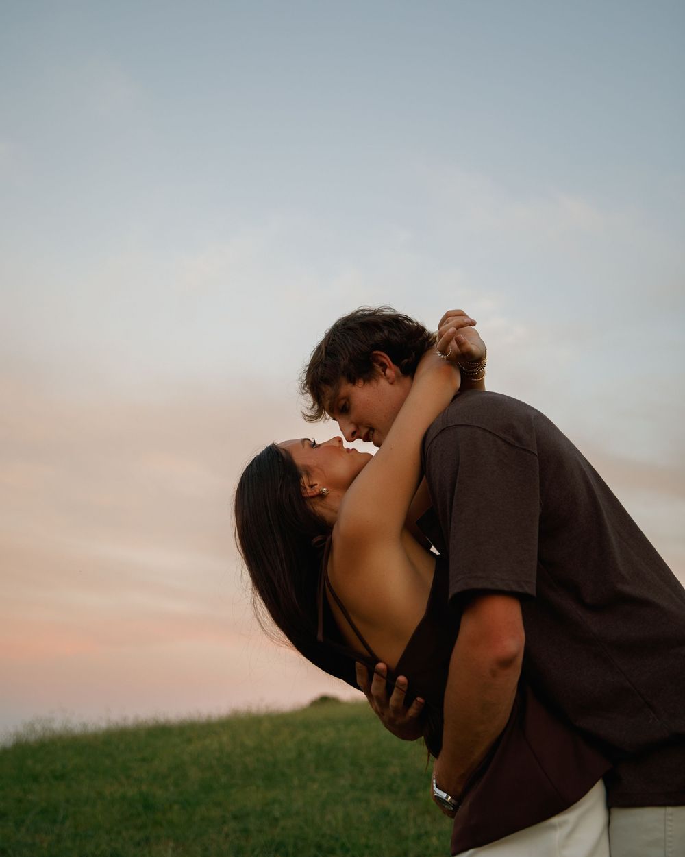 Couple sharing a romantic dip kiss at sunset on a grassy hill with a pastel sky backdrop.