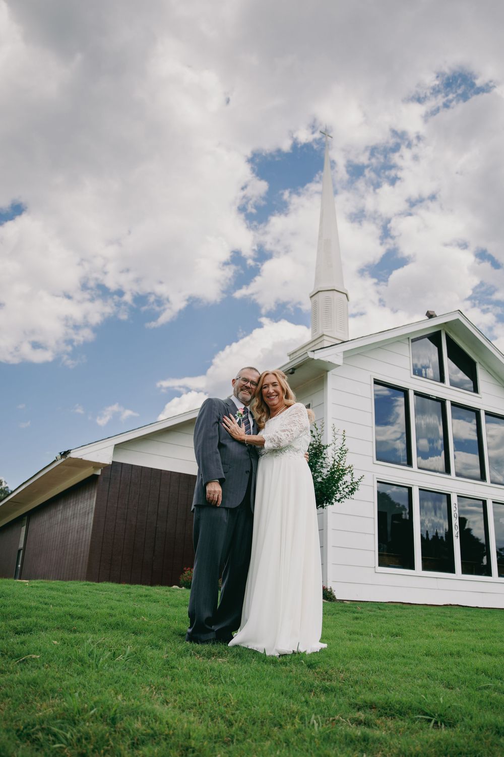Bride and groom embracing on green lawn in front of white chapel with steeple under cloudy blue sky.