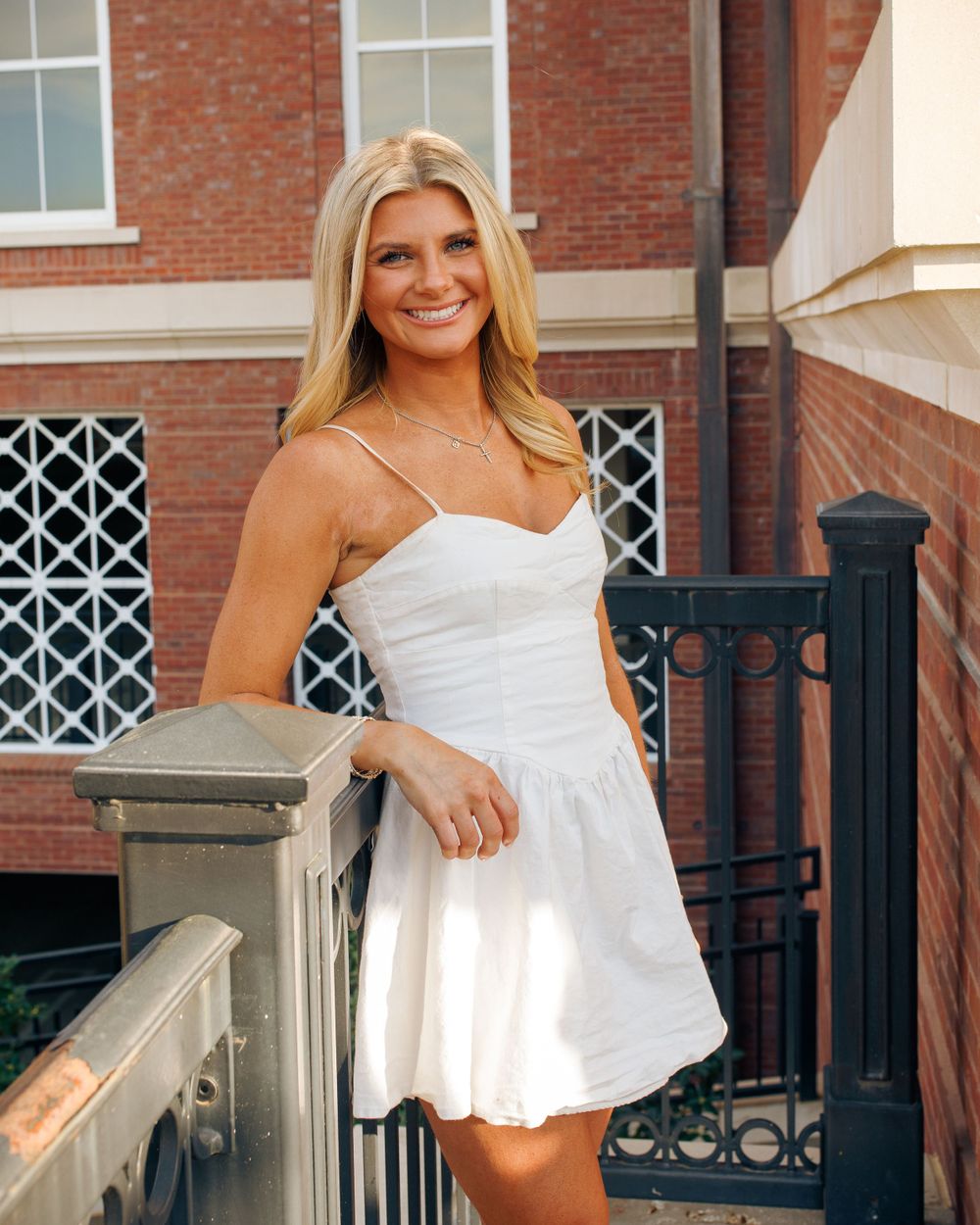A person in a white sundress stands smiling by a brick building entrance with ornate iron railings.