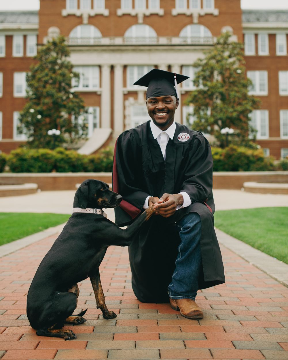 A smiling graduate in cap and gown kneels with a black dog on a brick path outside a university building.