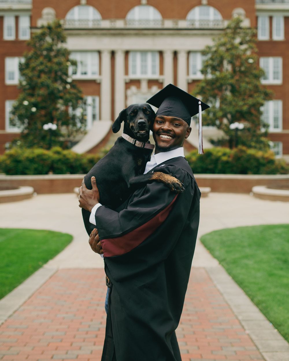 A smiling graduate in cap and gown holds a black dog outside a brick university building on graduation day.