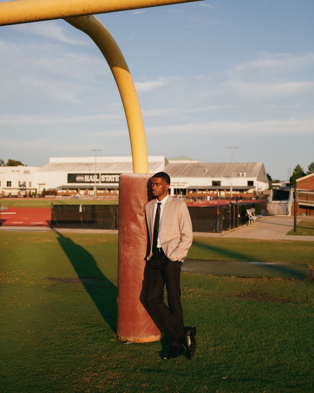 Young man in suit leaning against football goal post on green field at golden hour.
