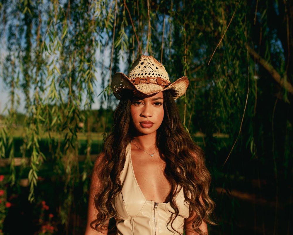 Young woman in a straw cowboy hat and white dress poses under a weeping willow tree at golden hour.
