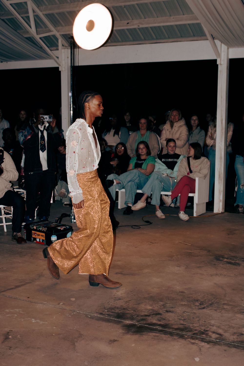Model walks the runway wearing a white top and gold pants under dramatic overhead lighting.