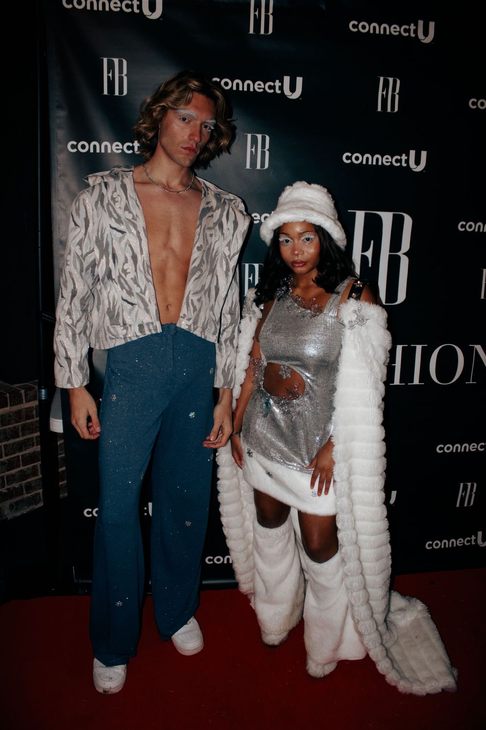 Two models pose on a red carpet at a fashion show.