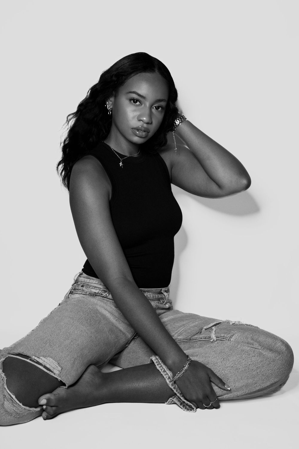 Model in black sleeveless top and distressed jeans poses against light background.