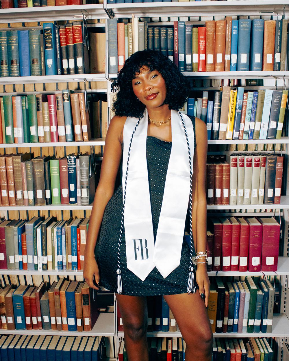 Graduate in dark dress and white stole poses in front of library bookshelves.