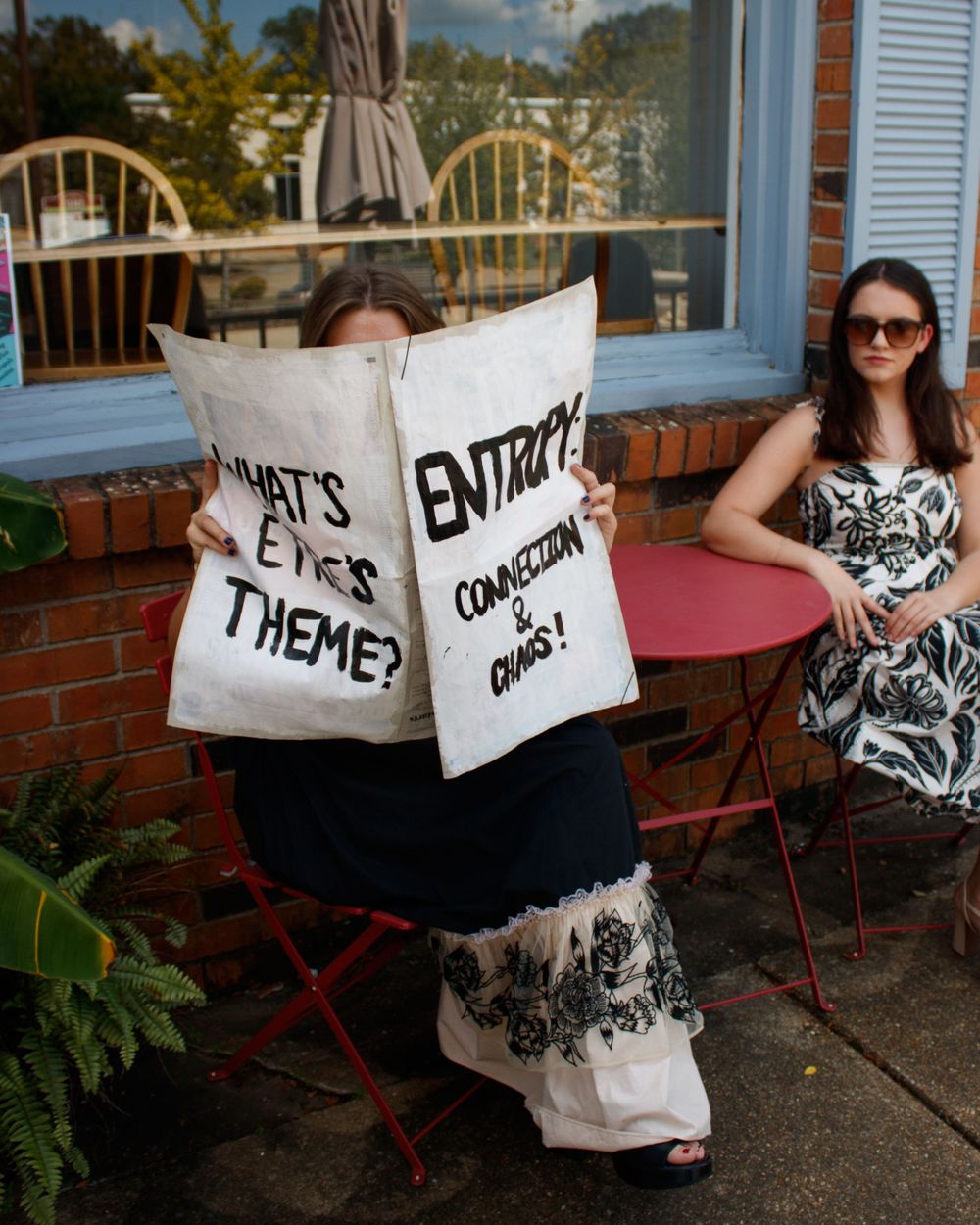 Two people posing with text signs on a brick terrace with outdoor furniture and plants.