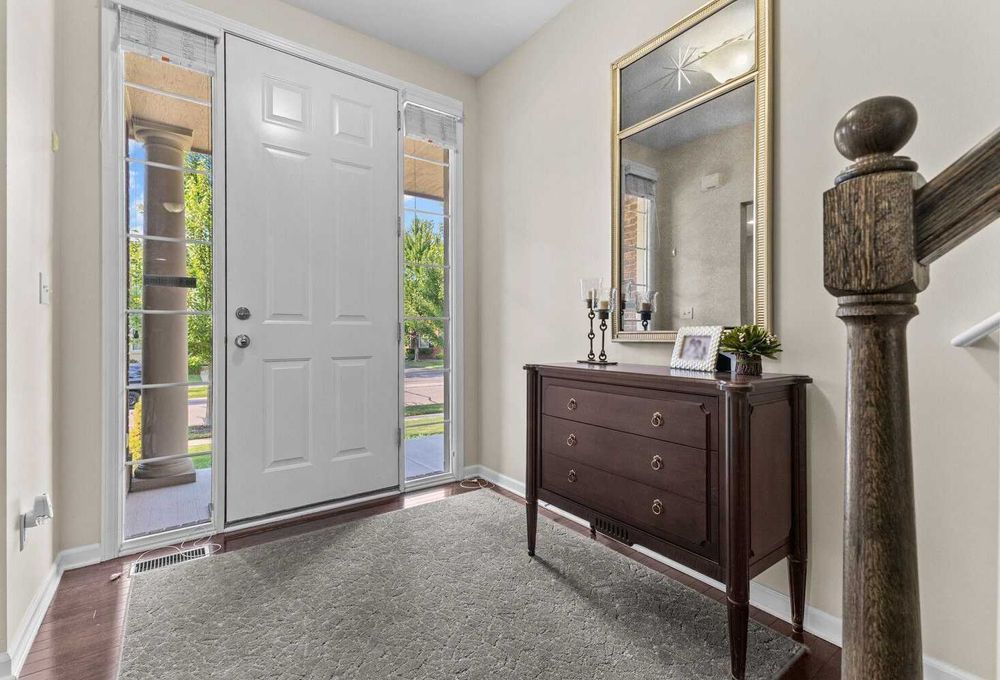 Bright entryway with white paneled door, dark wood dresser, gold mirror, and carpeted staircase.