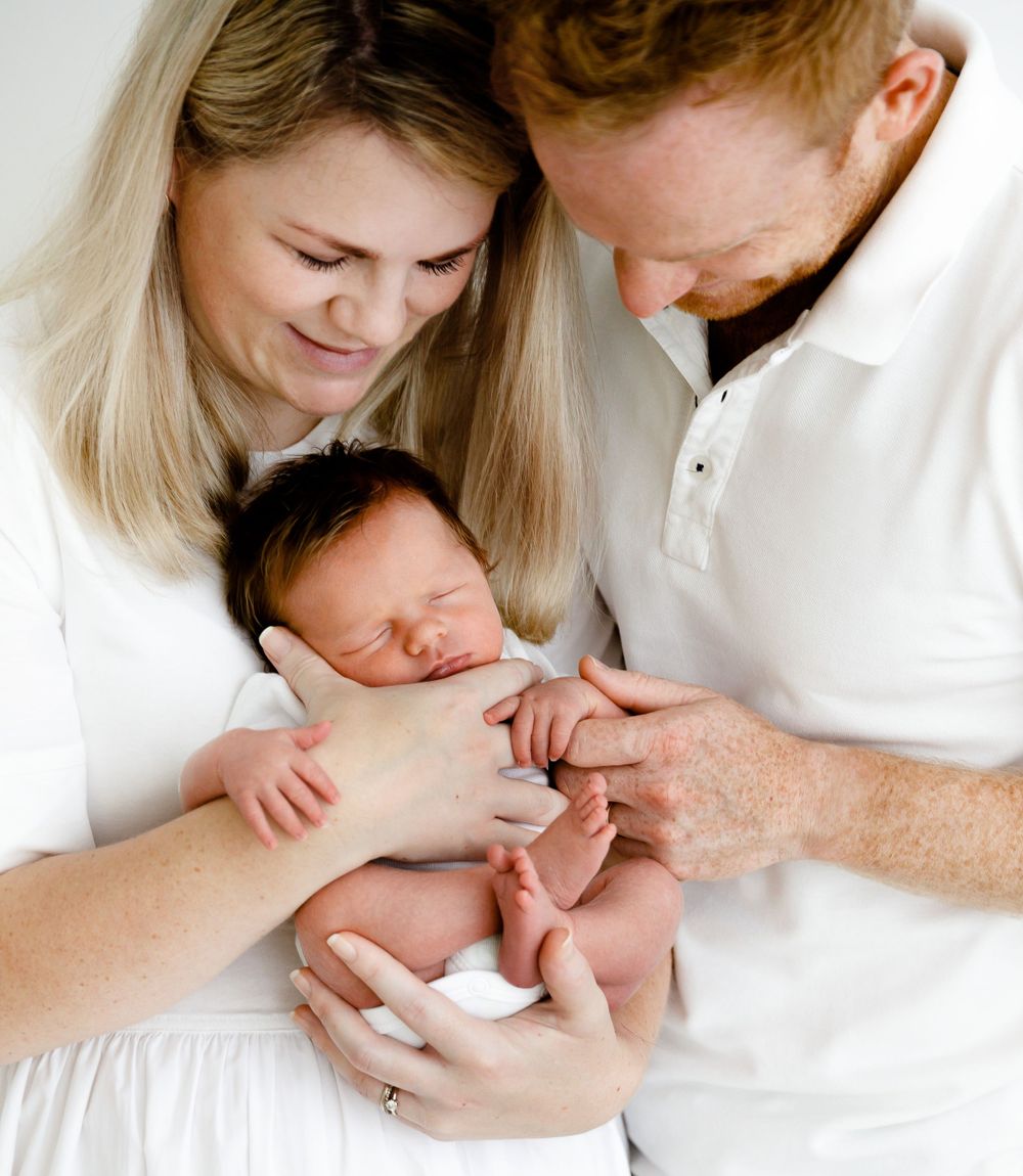 Parents in white clothing tenderly holding their sleeping newborn baby.