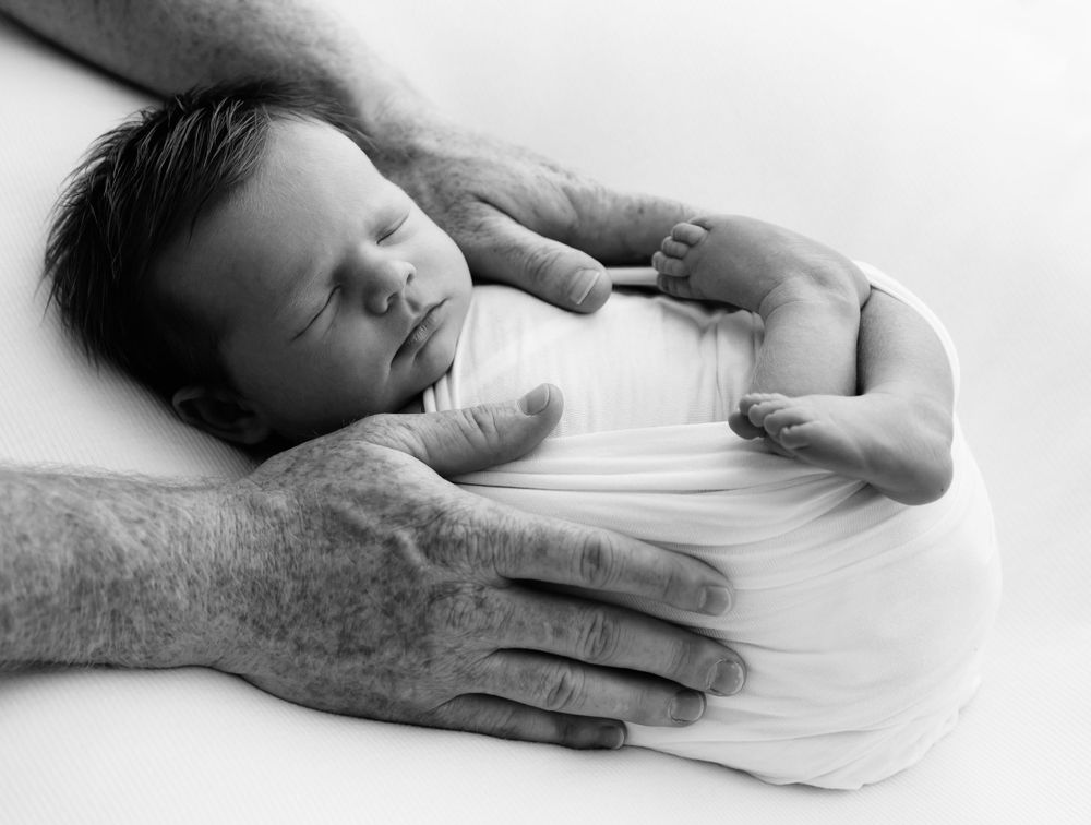 Black and white image of strong hands gently cradling a sleeping newborn wrapped in white.