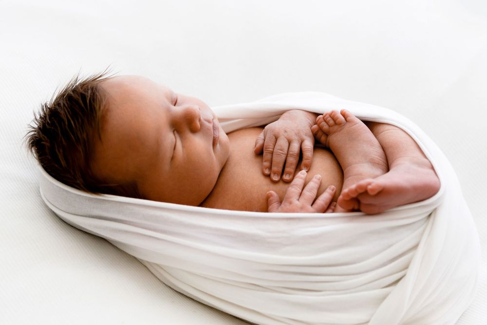Peaceful newborn baby wrapped snugly in white blanket while sleeping on light background.
