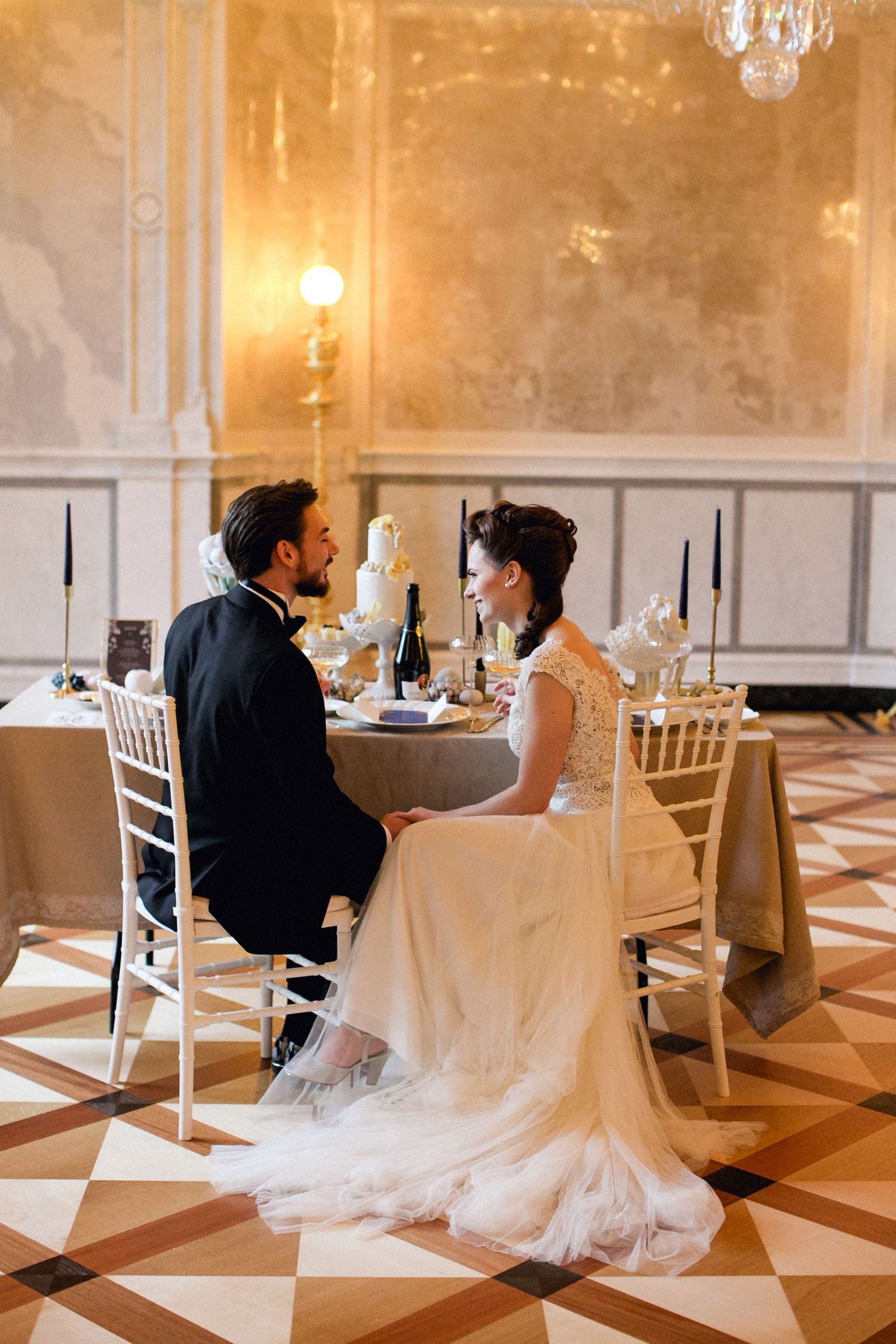 Elegant wedding couple sits at a table in a luxurious ballroom with black and white checkered floors and crystal chandeliers.