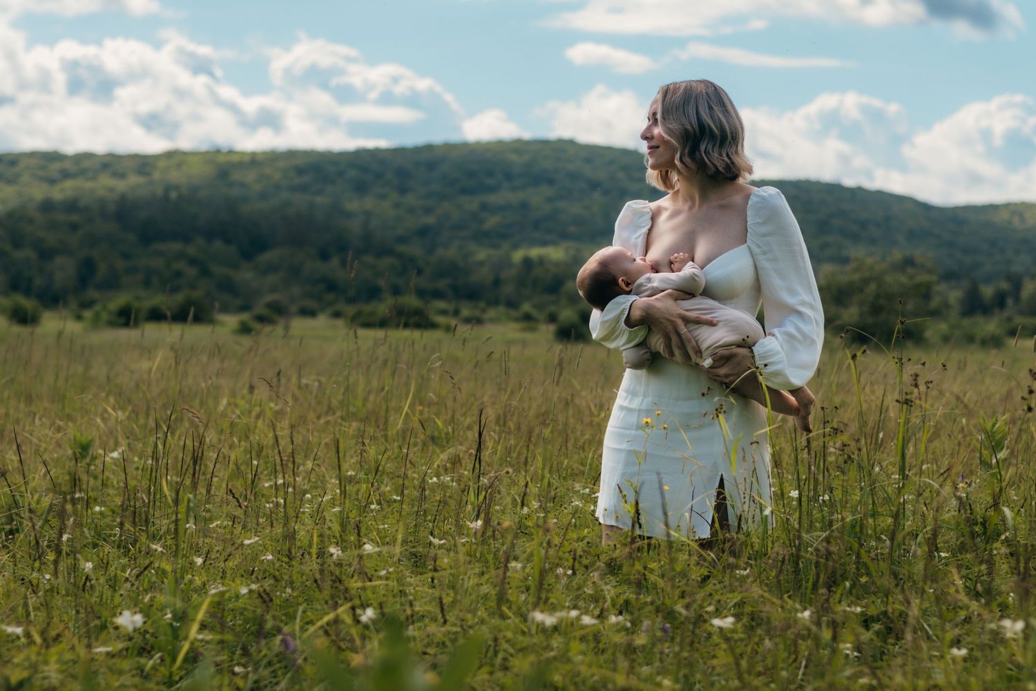 A mother standing breastfeeding her baby in a large grassy field.