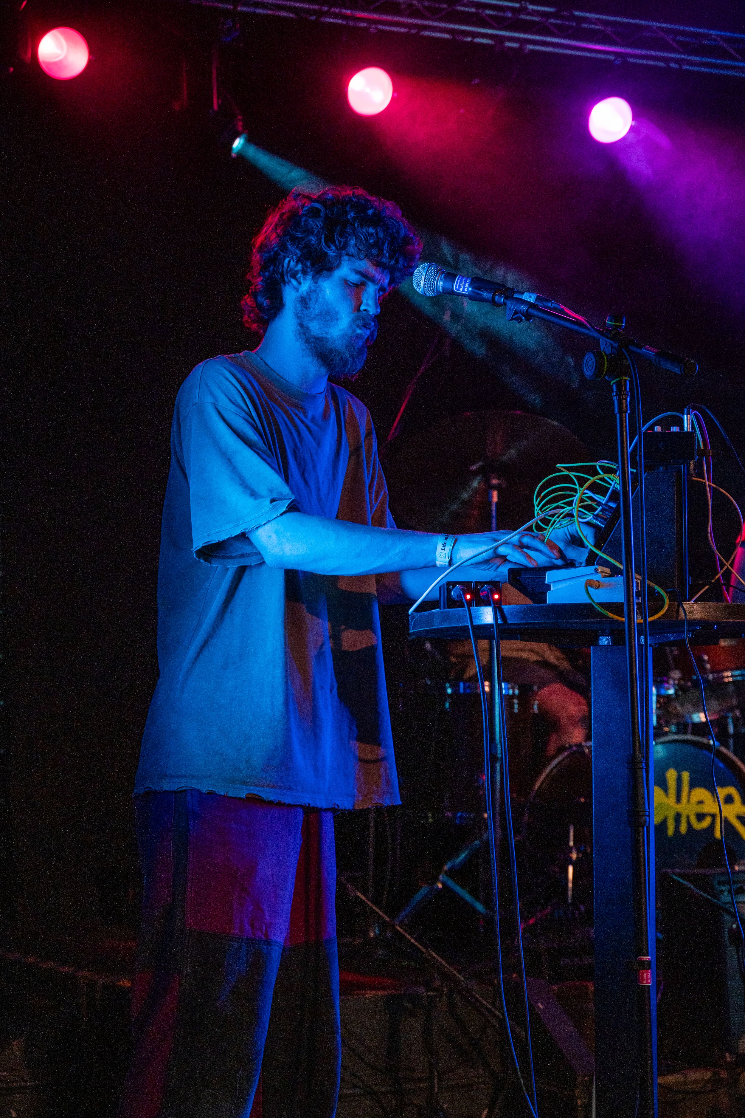 Musician performs at synthesizer keyboard station under dramatic purple and blue stage lighting.