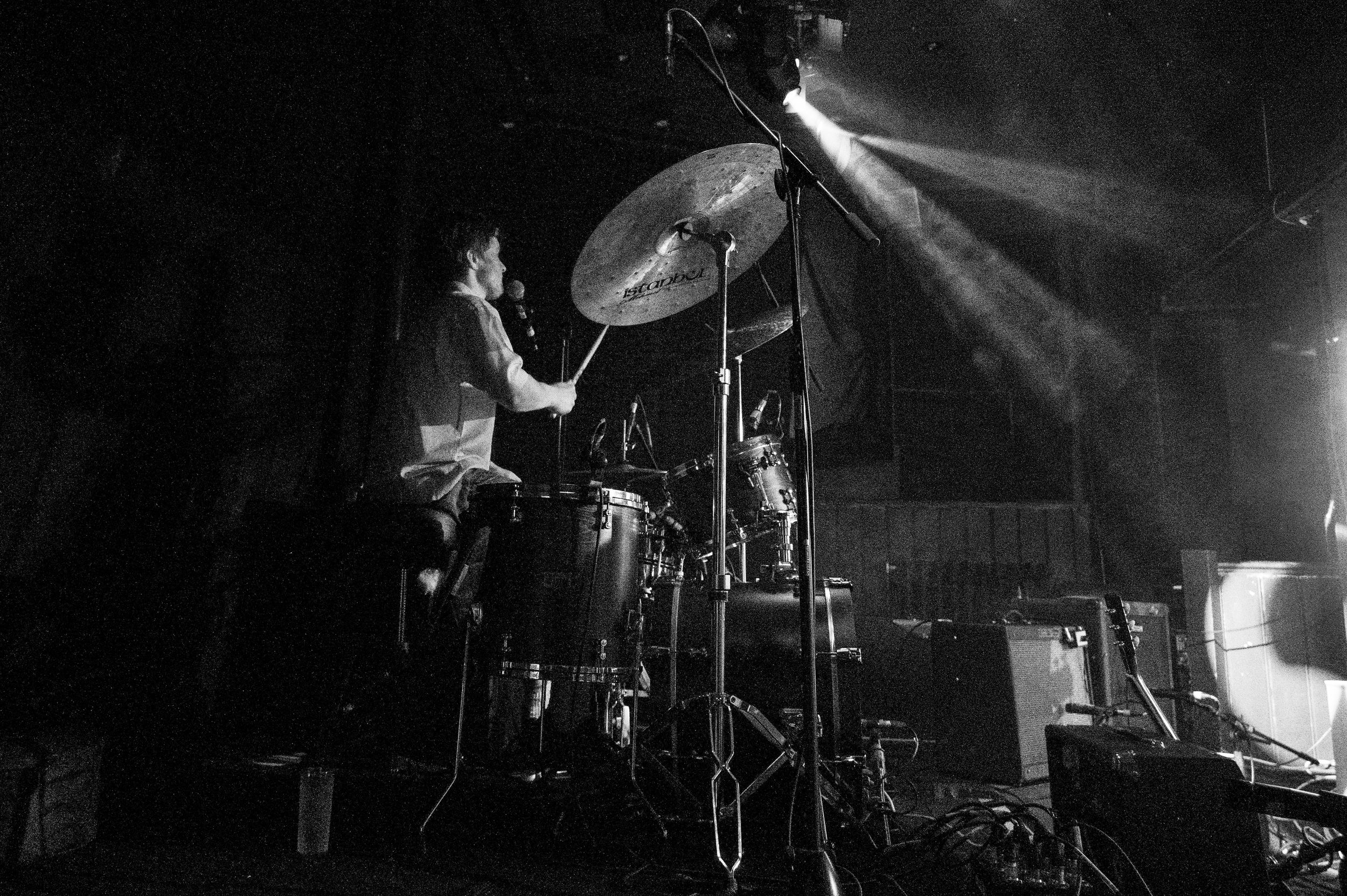 Drummer performing energetically on stage captured in dramatic black and white concert photography.