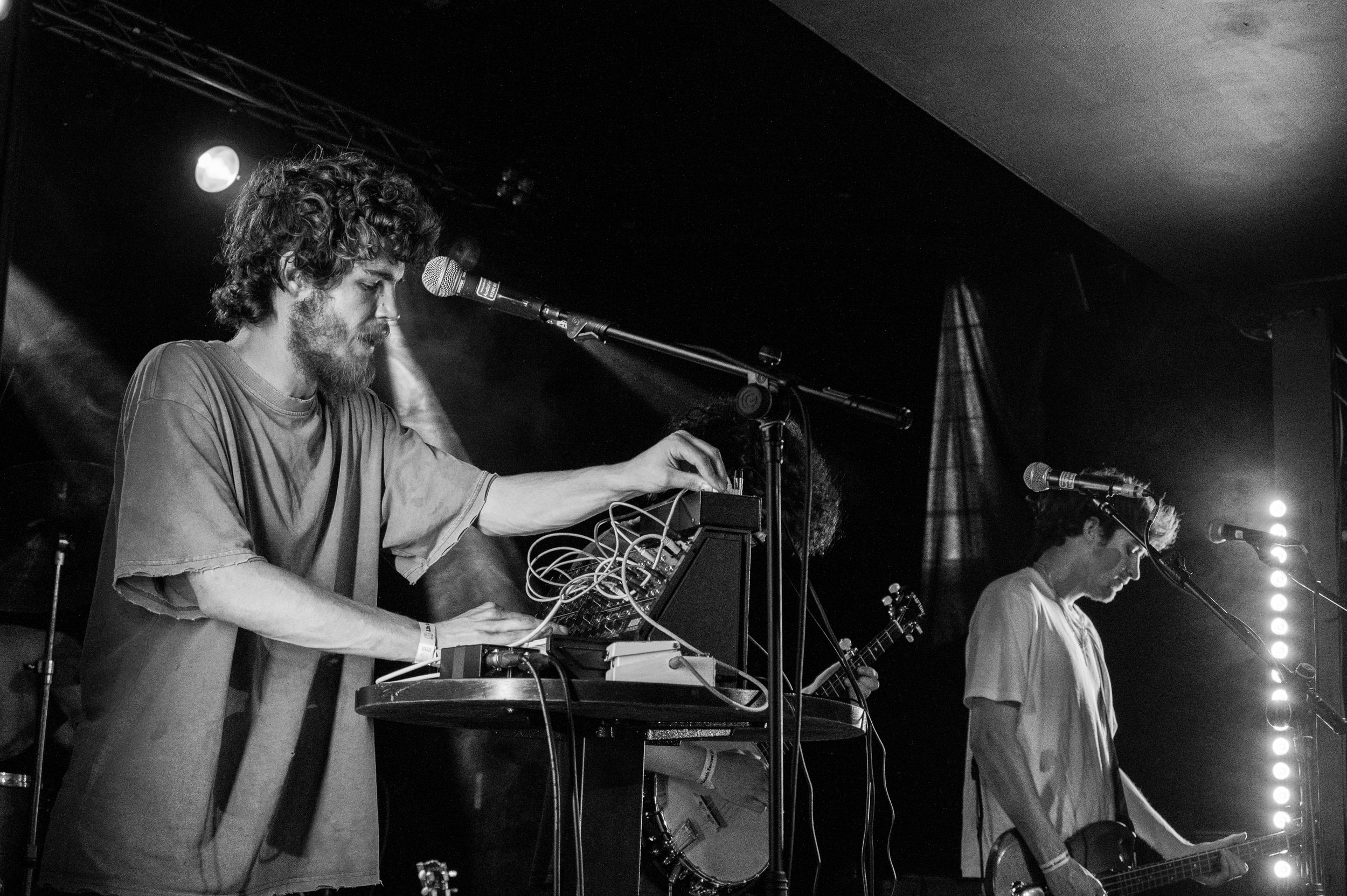 Musicians perform with electronic equipment under moody concert lighting in black and white.
