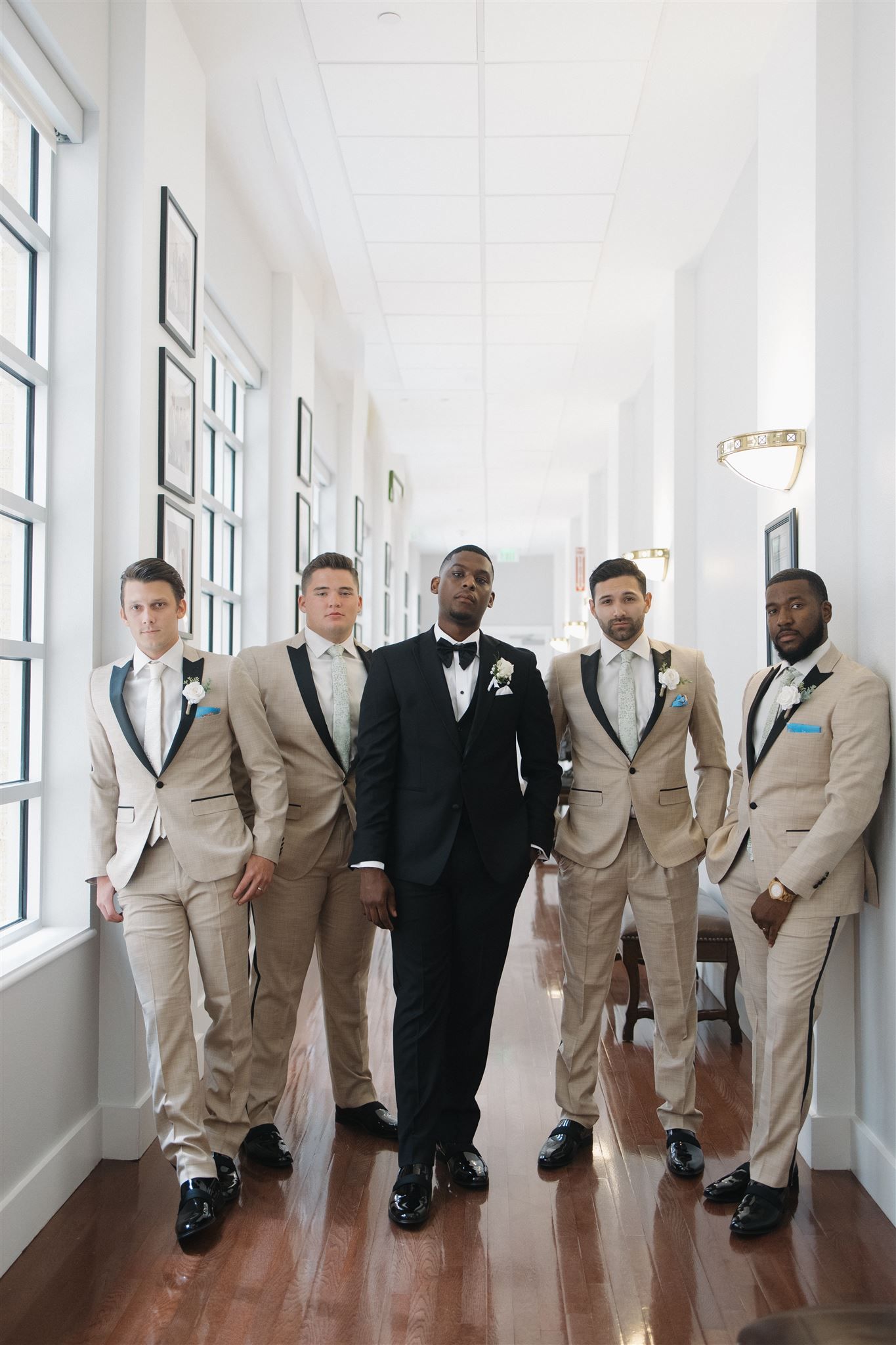 Groomsmen in coordinating tan suits stand with groom in black tuxedo in bright hallway with hardwood floors.