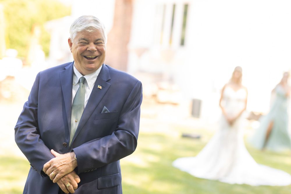Father of the bride laughing with joy during first look as bride stands in background in white gown.