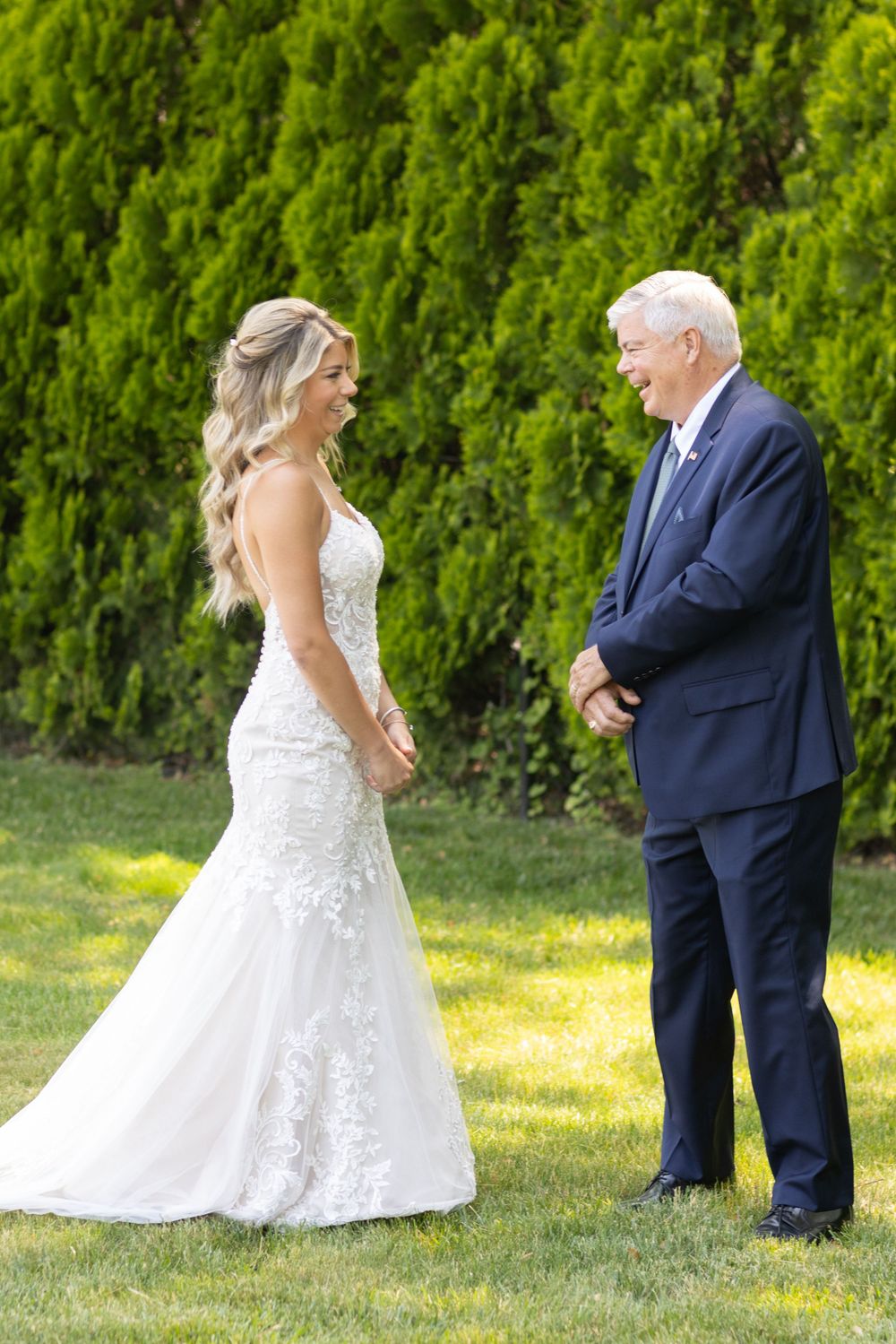 Bride in lace wedding gown smiling at older man in navy suit during outdoor first look ceremony.