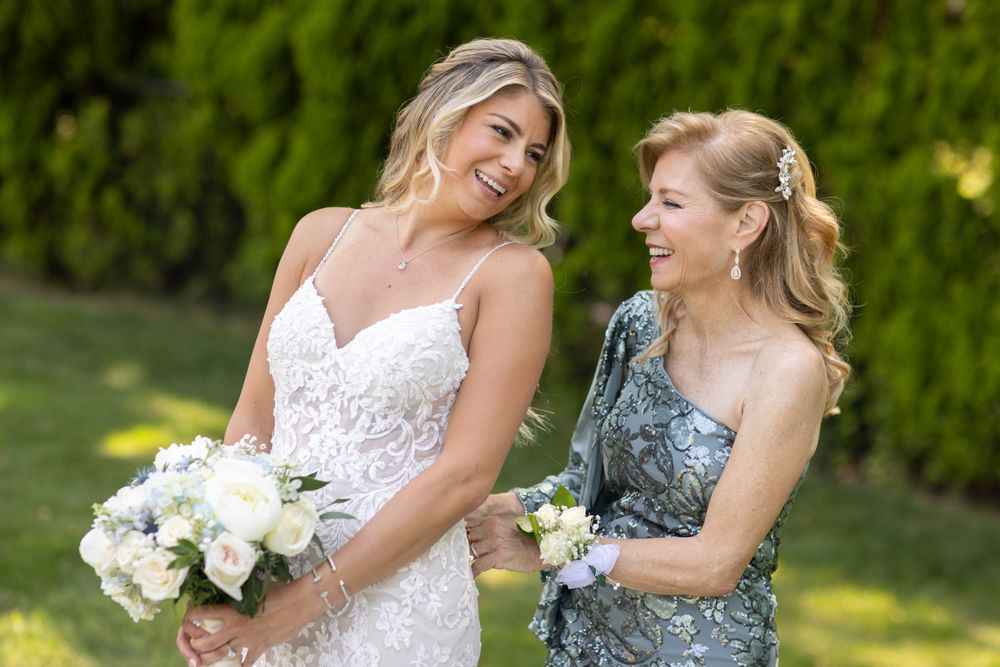 Smiling bride in lace gown holding white bouquet stands with mother in silver dress outdoors.