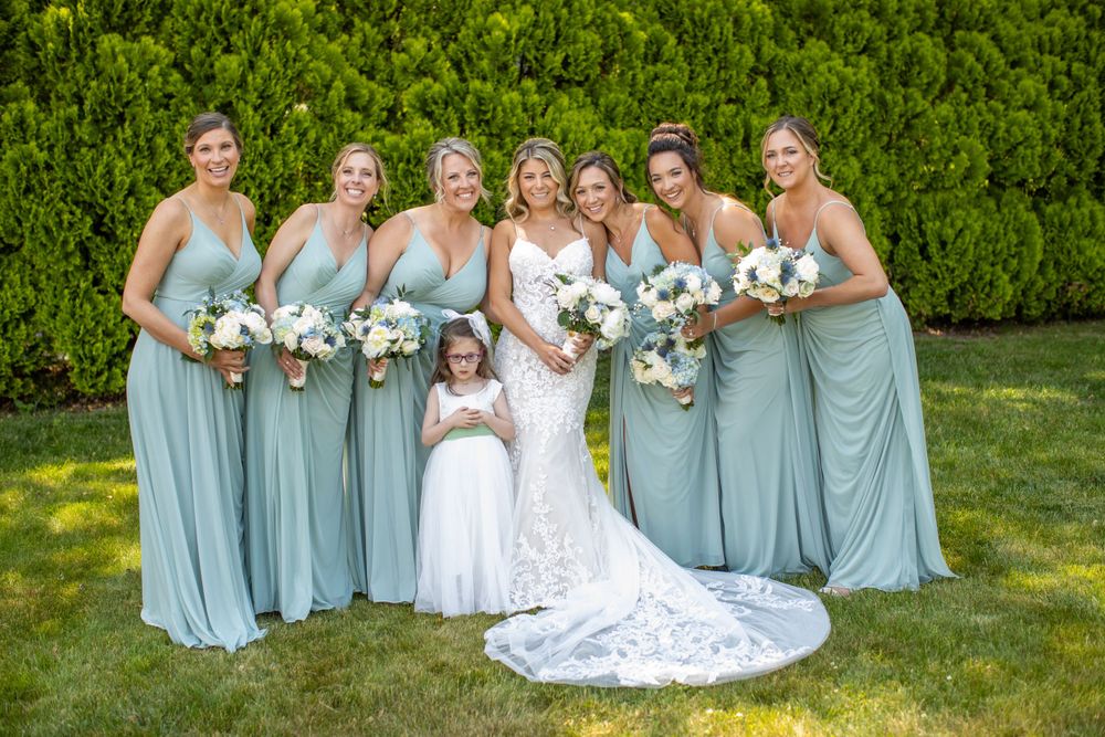 Bride in lace gown poses with six bridesmaids in sage green dresses and flower girl outdoors.