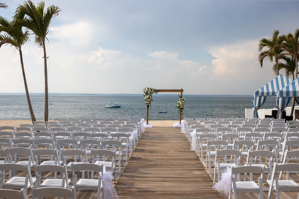 Elegant beachfront wedding ceremony setup with white chairs, wooden arch, and ocean backdrop.