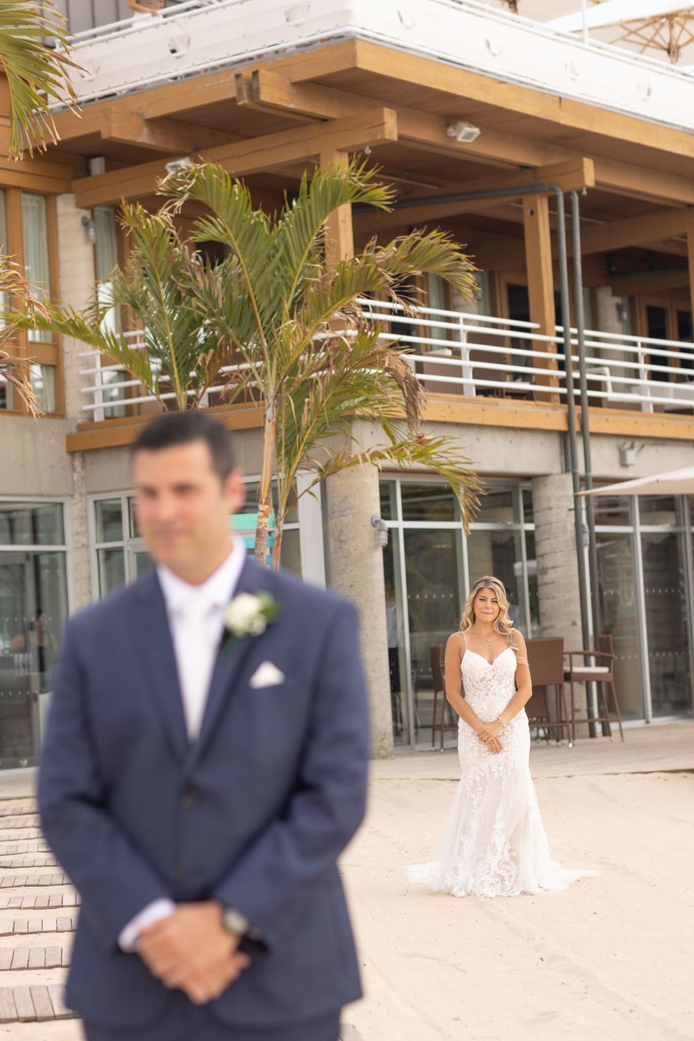 Bride in lace gown approaches groom in navy suit for first look outside modern beach venue.