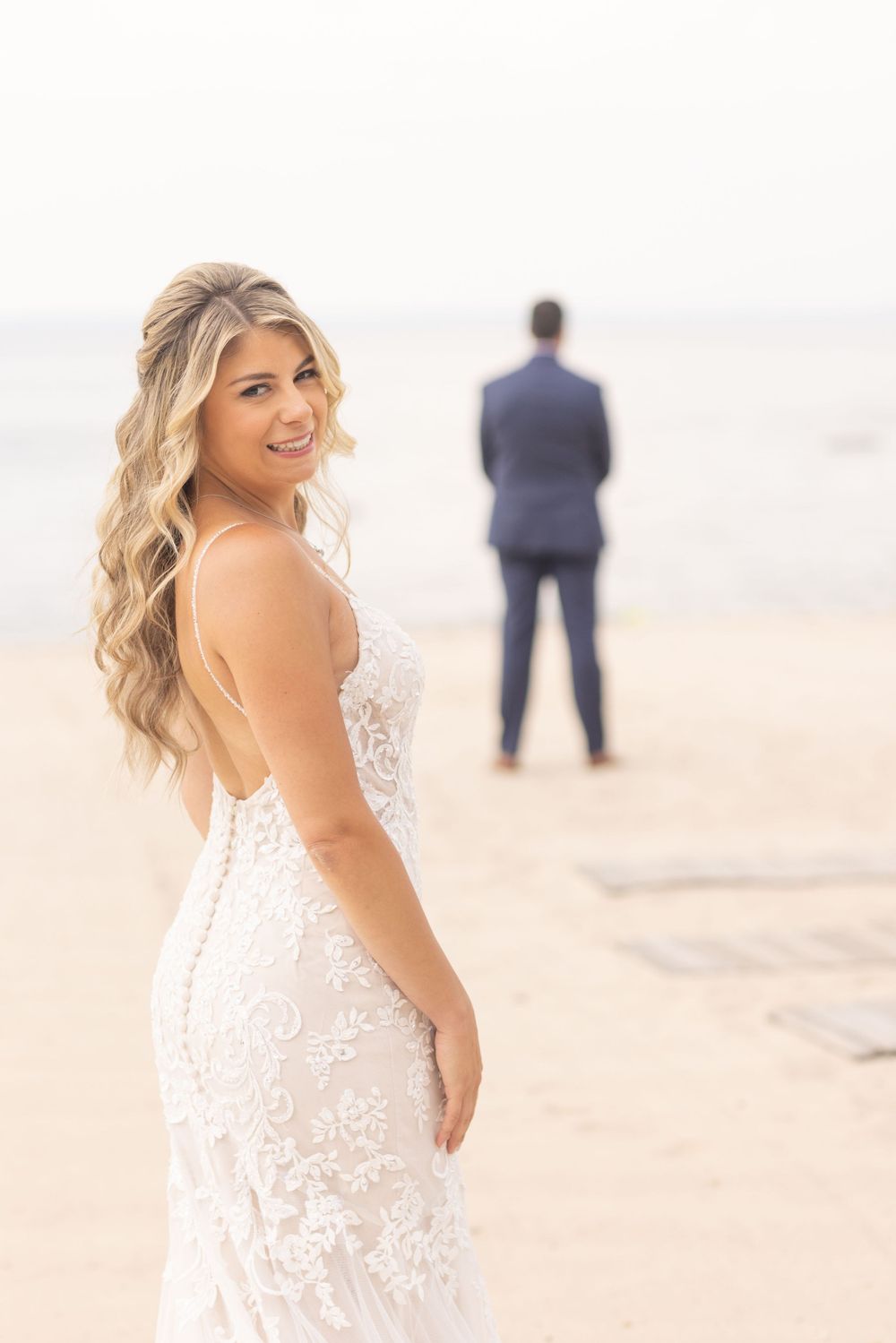 Radiant bride in lace gown smiles on beach with groom standing in background near water.