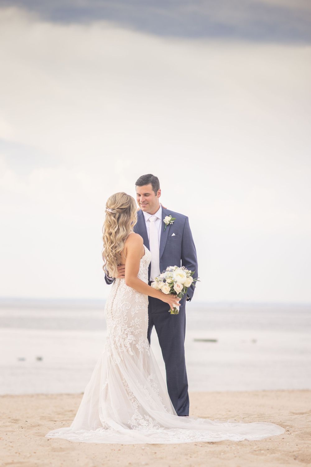 Bride and groom share romantic moment on sandy beach, groom holding white bridal bouquet.