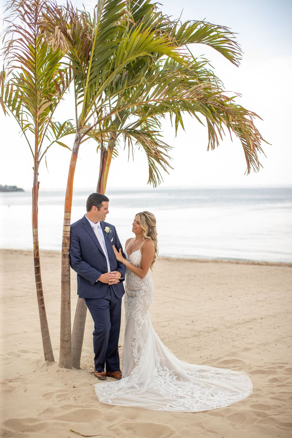 Bride and groom pose together under palm trees on sandy beach with ocean in background.