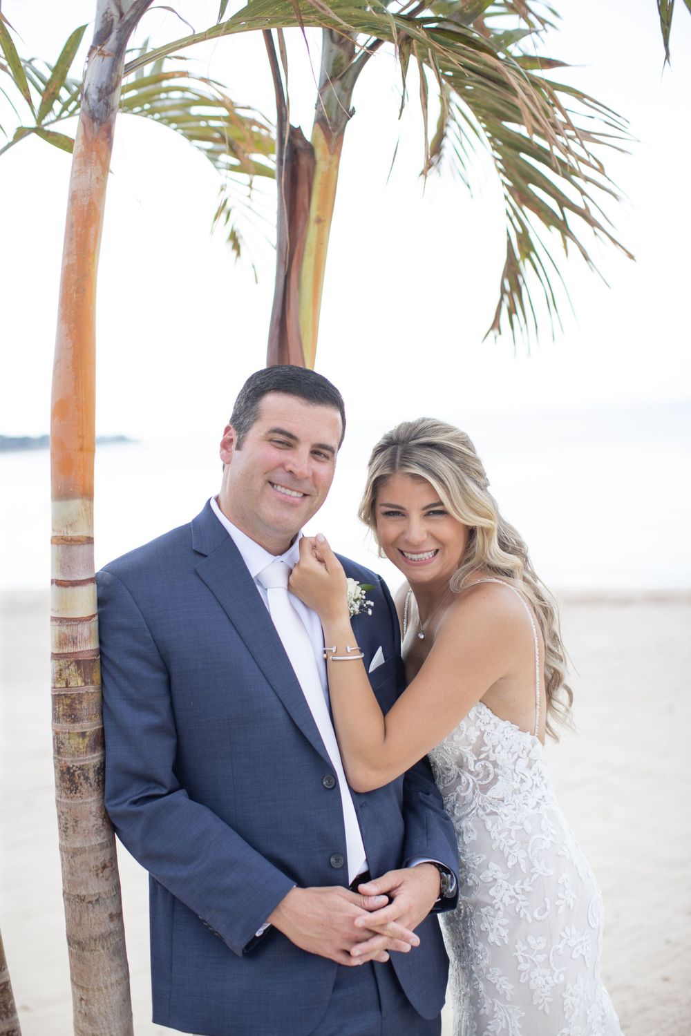 Smiling bride and groom pose together under palm tree on beach in wedding attire.