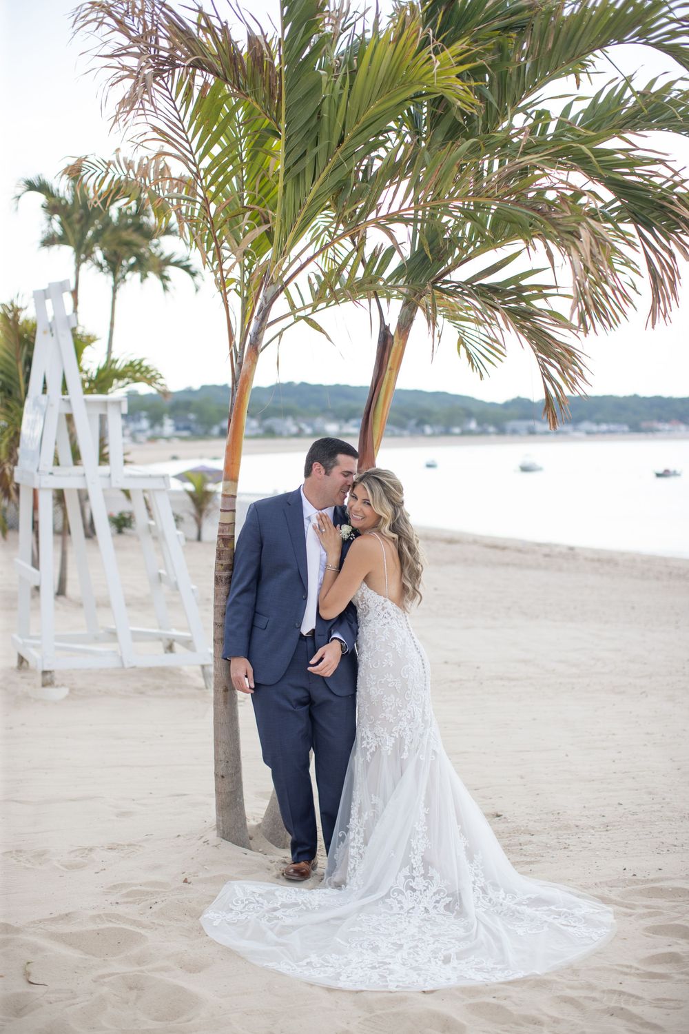 Bride and groom pose romantically under a palm tree on a sandy beach with ocean views.