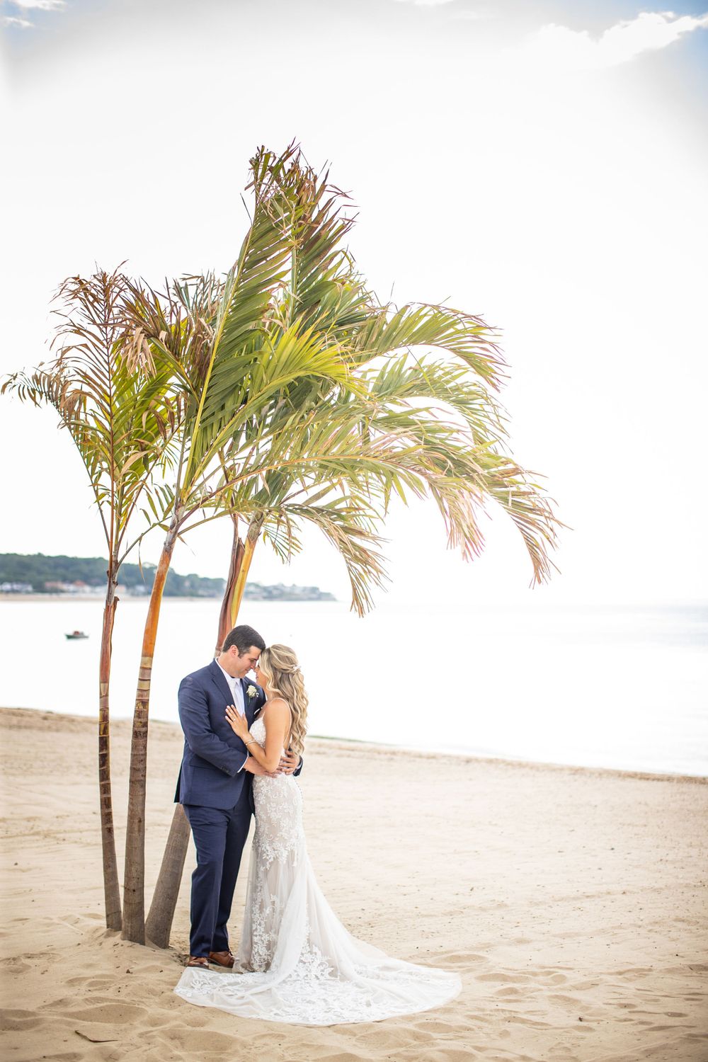 Bride and groom share a tender kiss under palm trees on a sunlit beach.