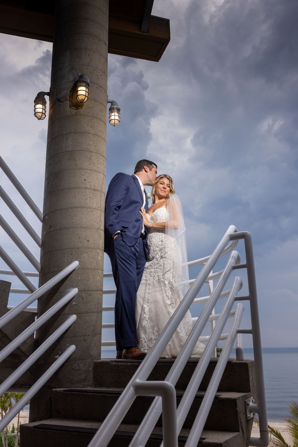 Groom in navy suit leans in to kiss bride in white lace gown on steps overlooking the ocean at sunset.
