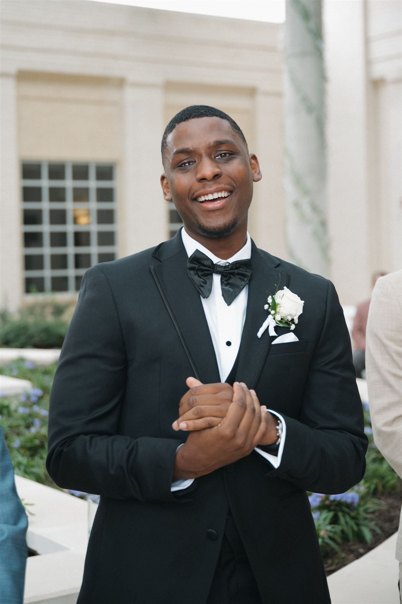 A groom in a classic black tuxedo and bow tie smiles while wearing a white boutonniere at an elegant venue.