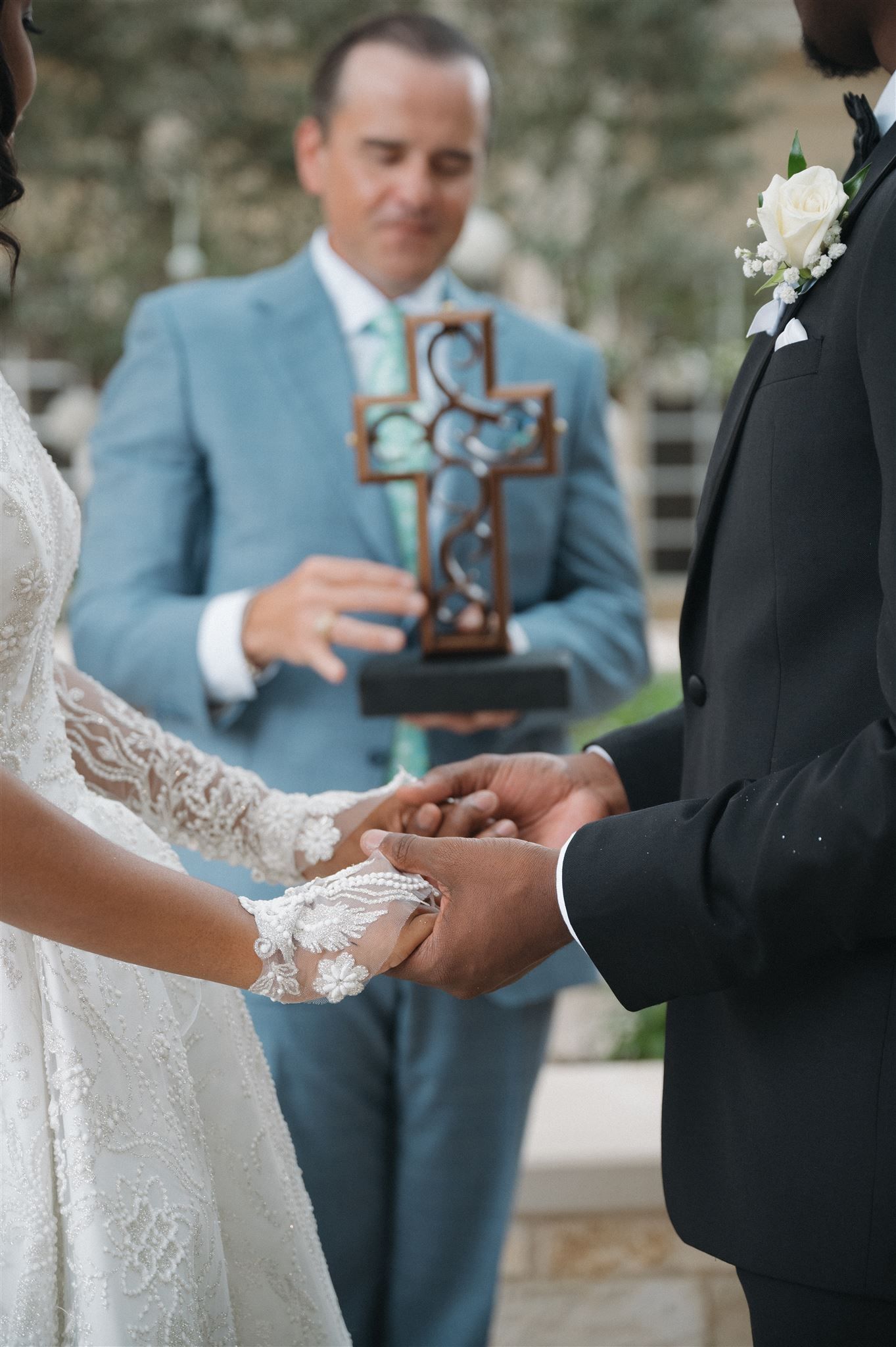 Close-up of bride and groom holding hands during wedding ceremony with officiant holding decorative cross in background.