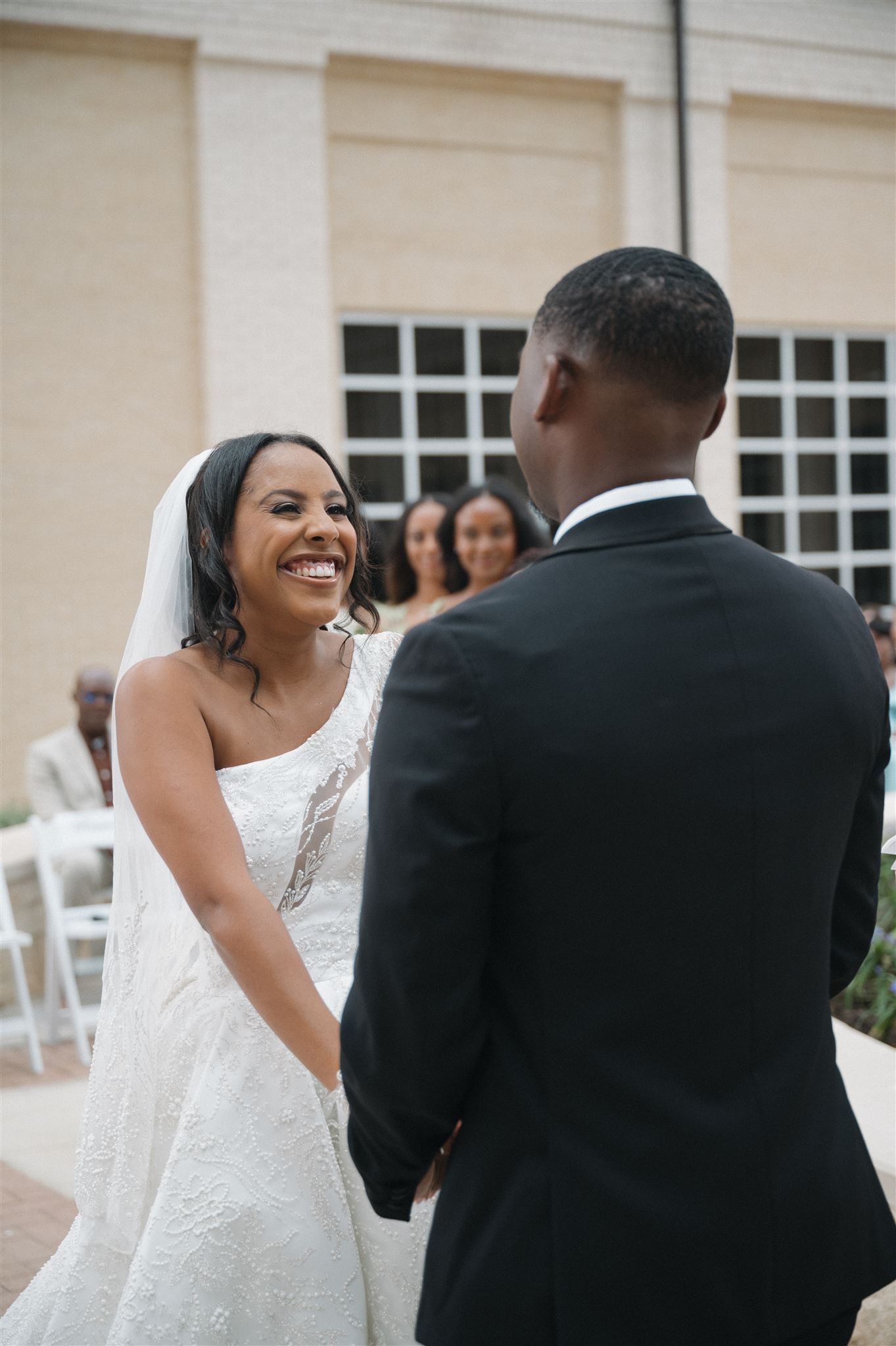 A joyful bride in a white lace wedding dress shares a happy moment with her groom during their outdoor ceremony.