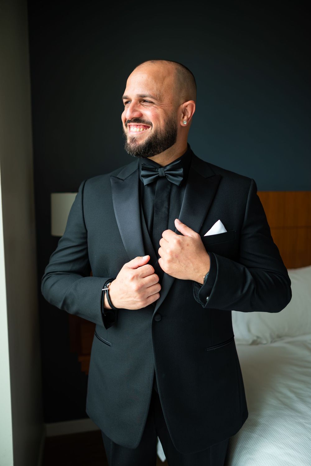 Groom in classic black tuxedo and bowtie smiling in hotel room.