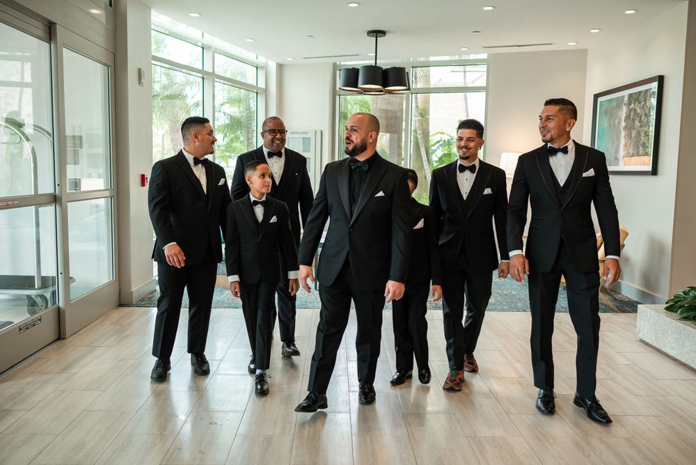 Groom walking with groomsmen in matching black tuxedos through modern hallway.