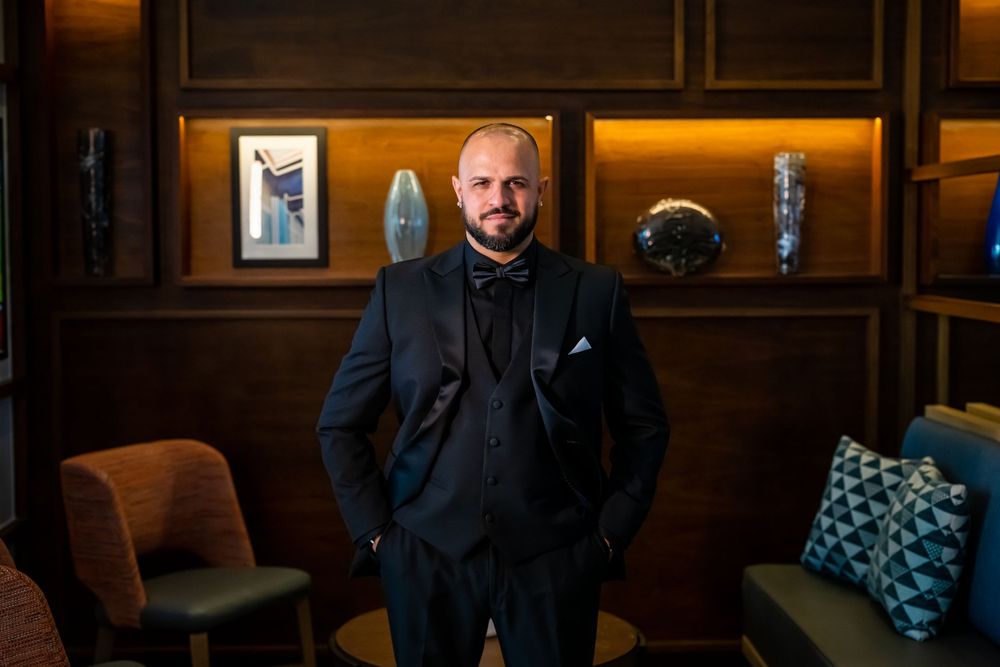 Groom in black tuxedo standing in front of wood-paneled wall with decorative shelving.